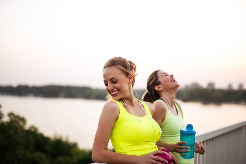Two women in workout clothes smiling after exercise by the water, holding water bottles