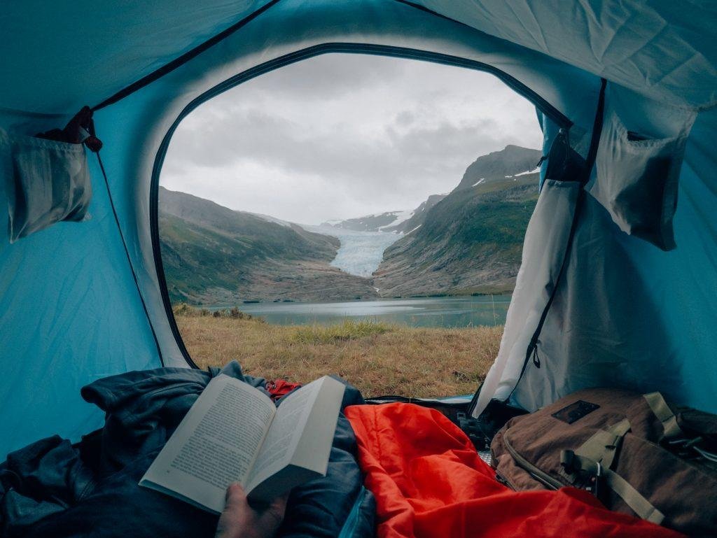 View from inside a tent of a glacier valley and lake, with a person reading a book in the foreground