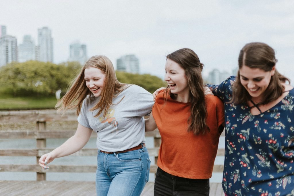 Three women walking arm in arm and laughing on a waterfront boardwalk with a city skyline in the background
