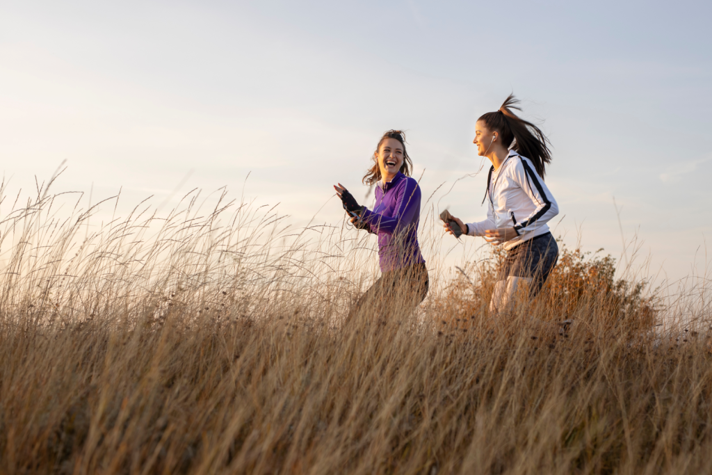 Two women jogging and laughing through tall grass at sunset