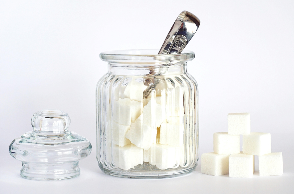 Glass jar filled with sugar cubes and a sugar tongs, with loose sugar cubes beside it