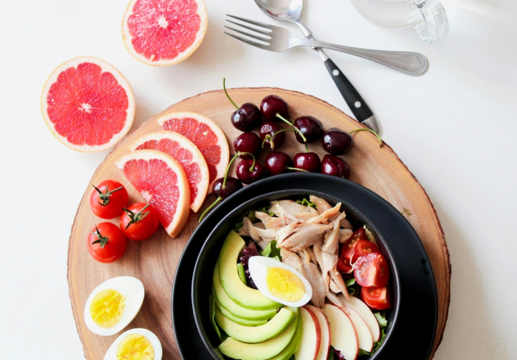 A salad bowl with avocado, chicken, egg, tomatoes, and apple slices, surrounded by grapefruit, cherries, and halved boiled eggs