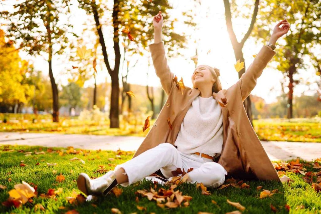 Woman sitting in a park with arms raised as autumn leaves fall around her